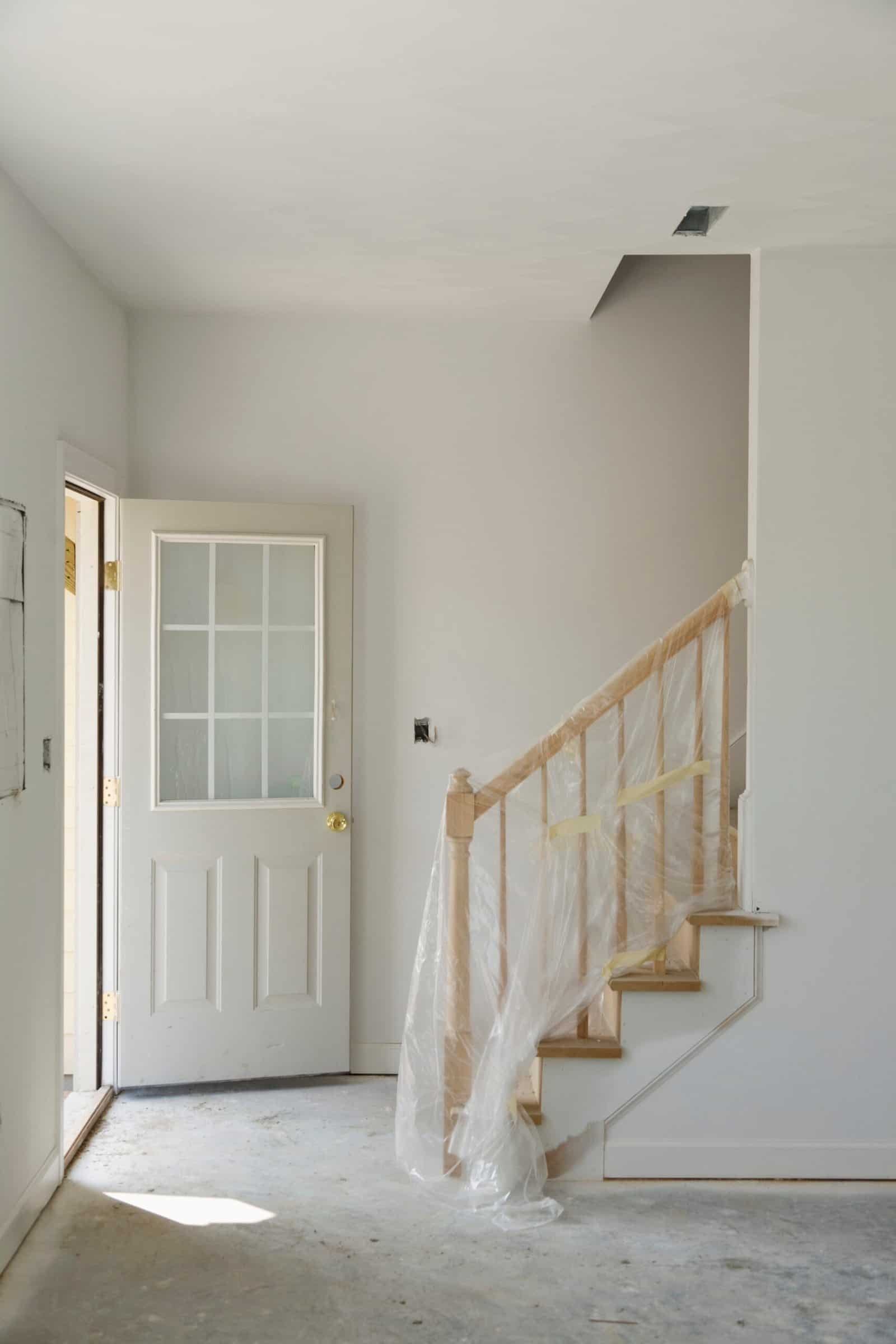 Freshly painted interior staircase area of a home under renovation, featuring a protective plastic cover on the banister. Bright white walls and a new door enhance the clean, modern look. Skilled local painters ensuring quality finish.